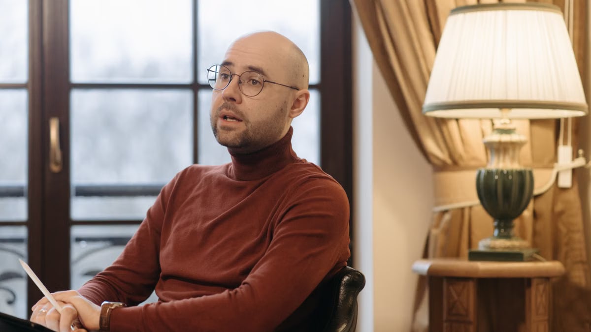Bald man in glasses speaking while seated indoors, with a warm lamp and curtains in the background.